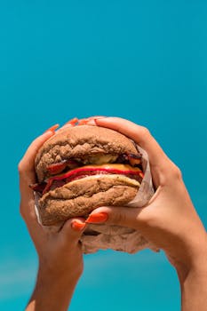Close-up of hands holding a tasty cheeseburger against a bright blue sky.