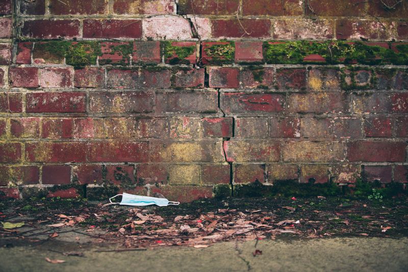 A face mask lays discarded against a moss-covered, cracked brick wall, symbolizing COVID-19 impact.