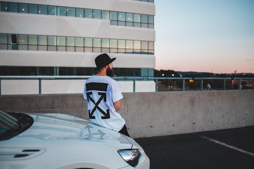 Fotografía de la vista trasera de un hombre anónimo elegante en una camiseta y sombrero elegante con gafas junto a un automóvil costoso cerca de un edificio contemporáneo