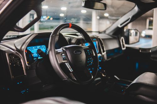 Free Black leather interior of modern automobile with steering wheel and glowing dashboard and instrument panel with blurred parking lot through windshield Stock Photo