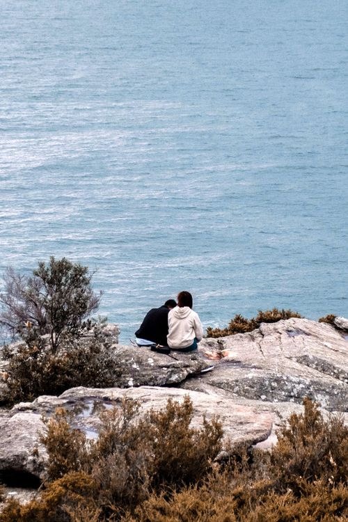 Free A couple sits on rocky cliffs overlooking the ocean, capturing a serene moment in Sydney, Australia. Stock Photo