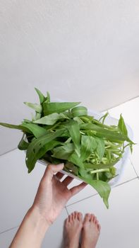 A person's hand holding fresh spinach leaves in a plastic tub with bare feet visible below.