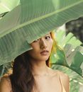 在阳光明媚的日子里，一位迷人的黑发亚洲女性站在覆盖部分脸的绿色异国叶子旁，注视着相机