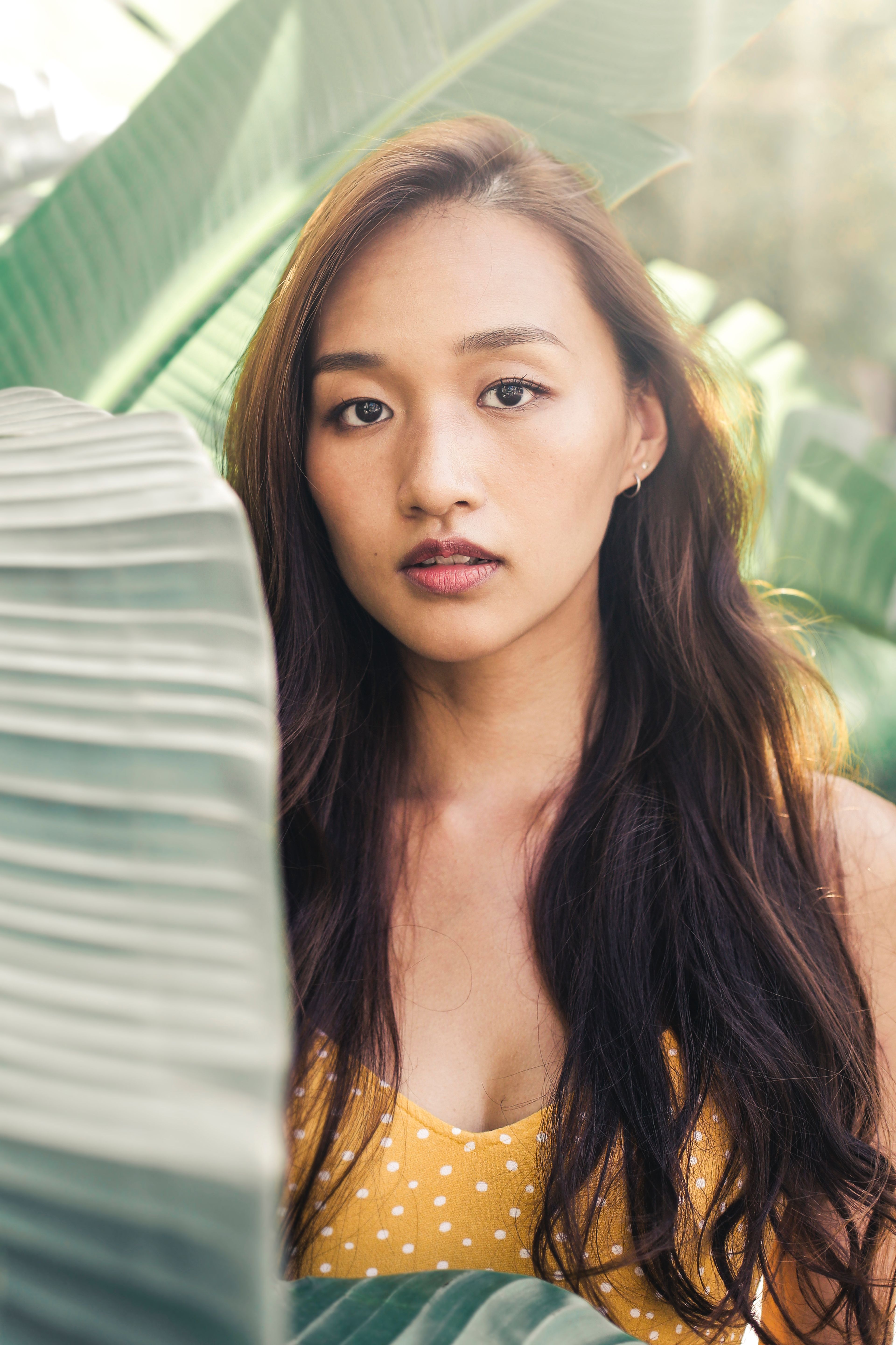 一名年轻女子身穿黄色圆点连衣裙，站在大片绿叶中，阳光洒落在她身上
