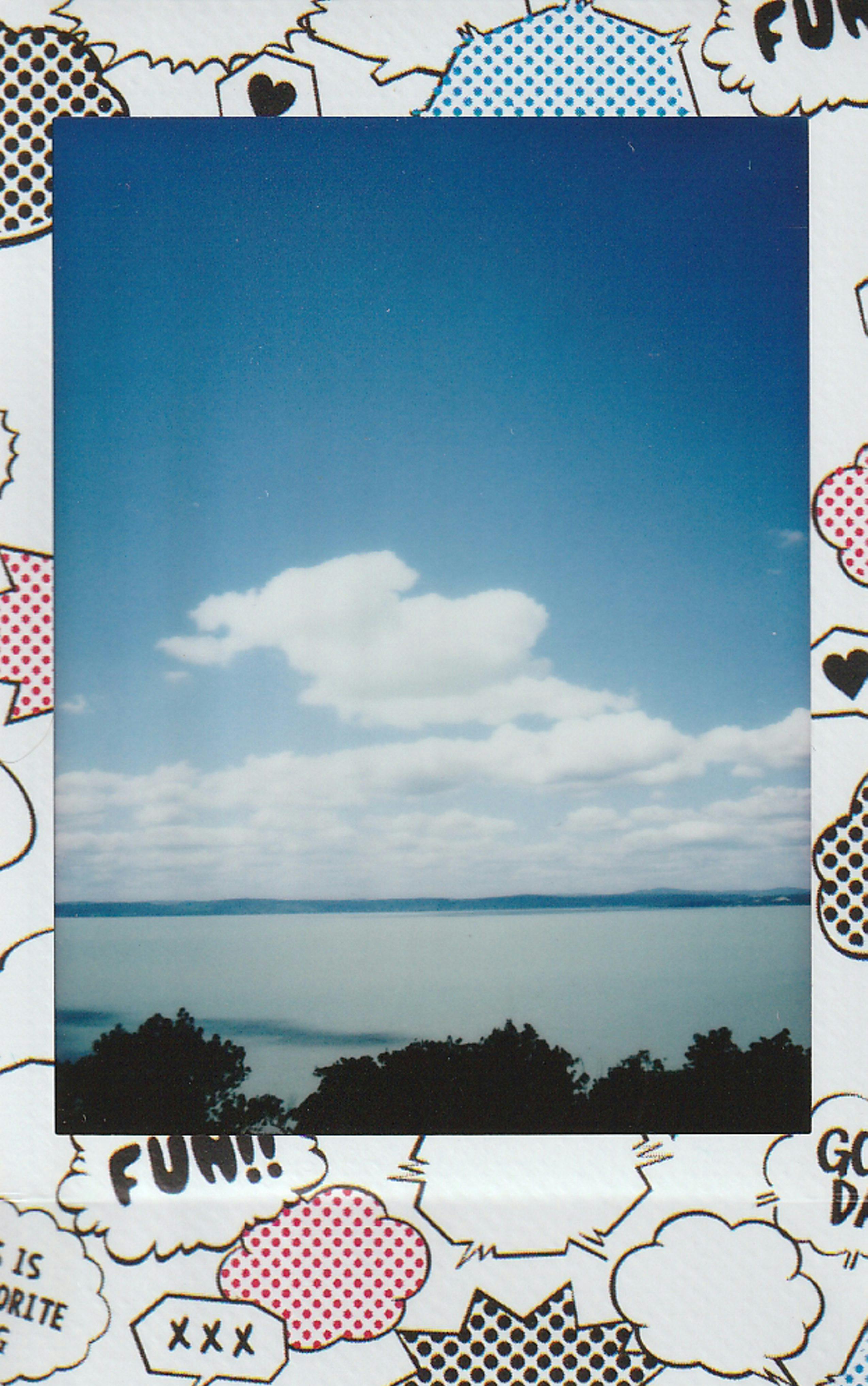 A serene lake view under a blue sky with fluffy clouds, captured on instant film.