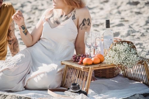 Free Relaxing beach picnic featuring wine, fresh fruits, and a camera set on a sandy beach. Stock Photo