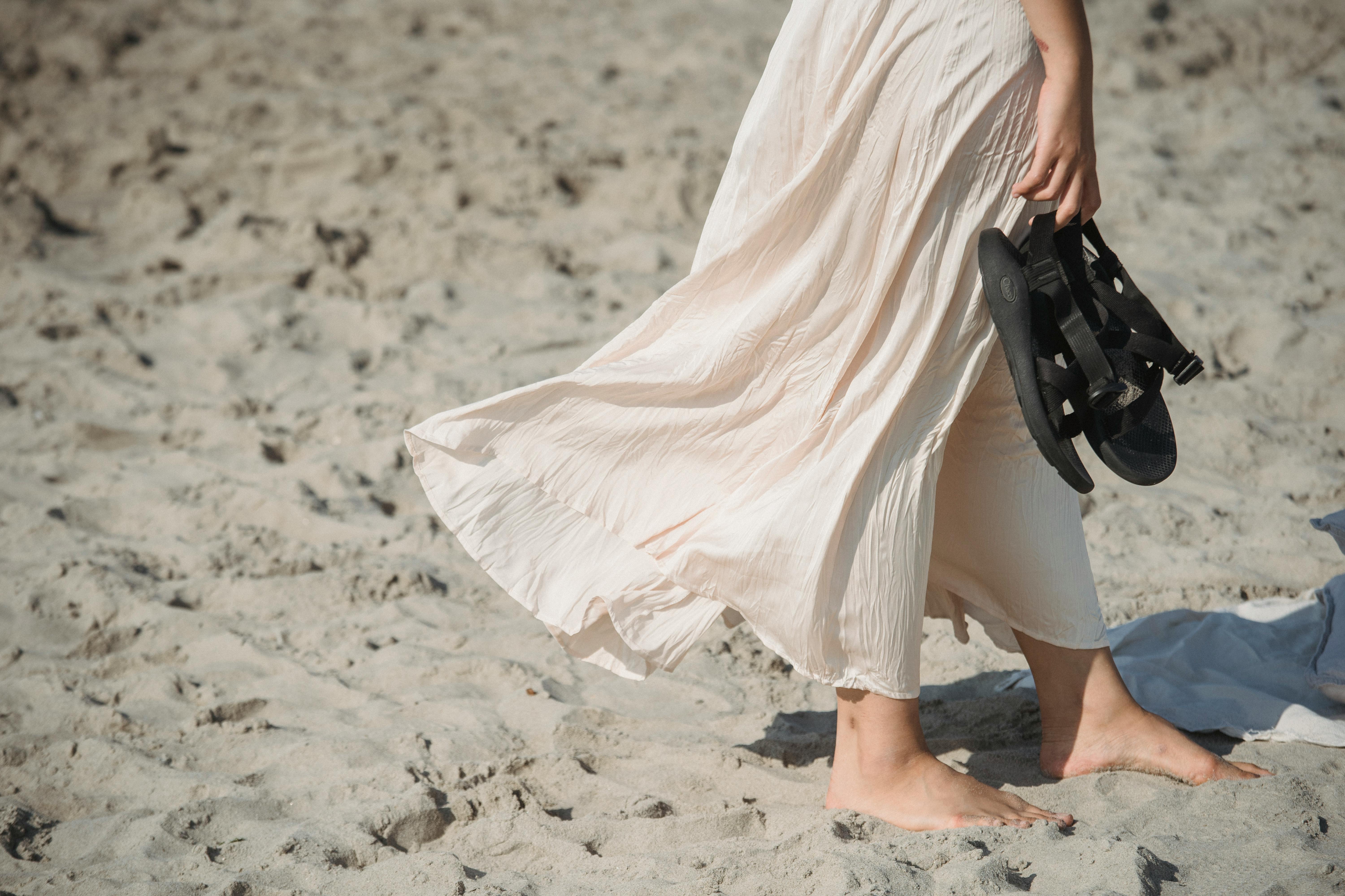 Free A woman strolls barefoot on a beach, holding her sandals with a breeze in her dress. Stock Photo