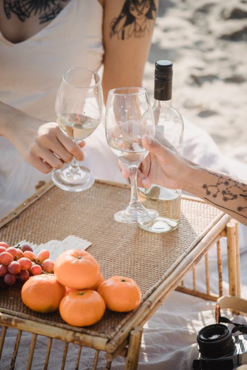 Free Two people toast with wine glasses at a beach picnic featuring grapes and oranges. Stock Photo