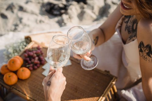 Free Beachside picnic scene with two people toasting with wine glasses amidst fruits on a sunny day. Stock Photo