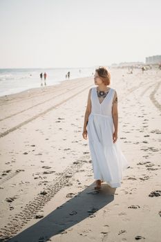 A woman in a white dress strolls along a sandy beach under clear skies, exuding tranquility.