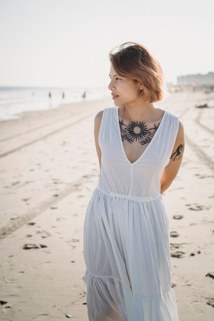 A young woman with tattoos walks on a sunny beach wearing a white dress.