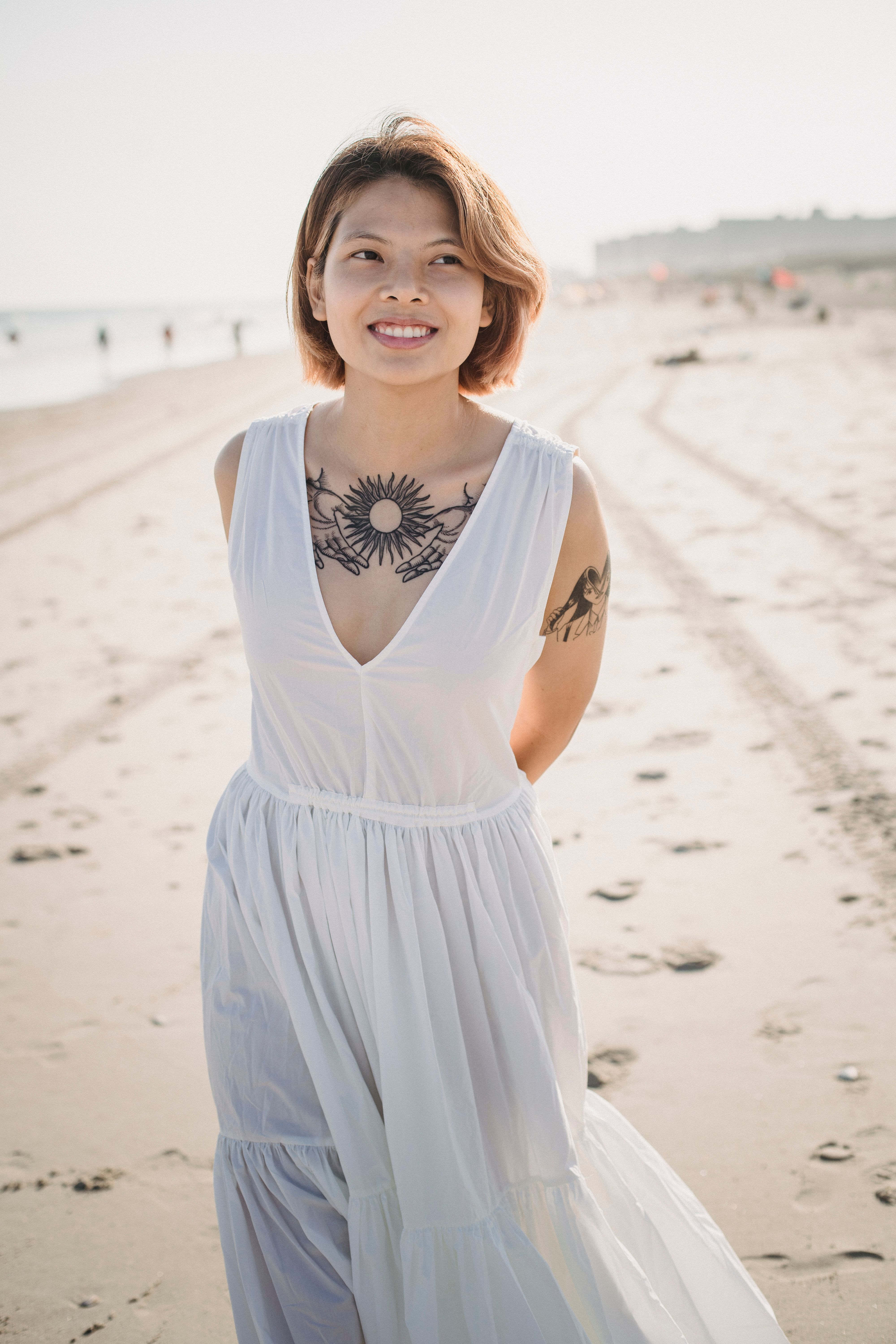 Smiling young woman with tattoos in a white dress enjoying a sunny day at the beach.