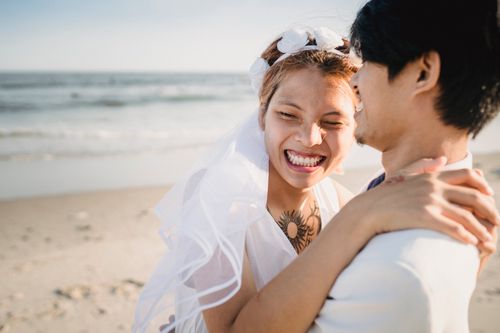 Free A laughing bride embraces the groom on a sunny beach, capturing a joyful wedding moment. Stock Photo