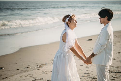 Woman in White Dress Holding Hands With Man in White Suit at Beach