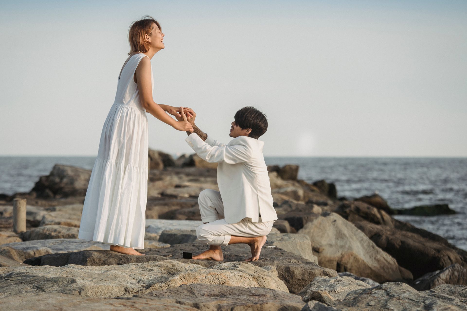 A couple shares a memorable marriage proposal by the sea on a rock-strewn beach.