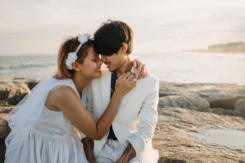 Free A couple in wedding attire embracing on a rocky beach at sunset, symbolizing love and romance. Stock Photo