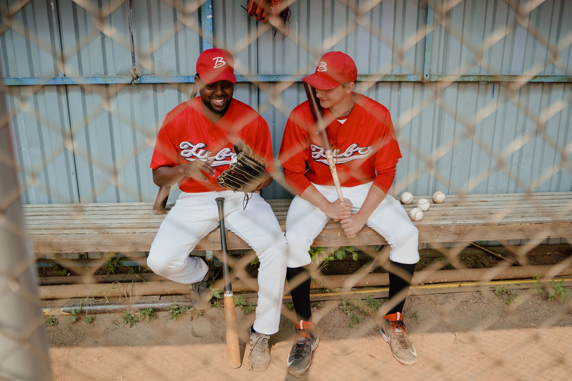 Two baseball players in red uniforms sitting on a dugout bench, chatting and smiling.