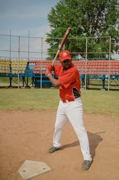 Black male baseball player in red uniform preparing to swing bat on a sunny day at a baseball field.