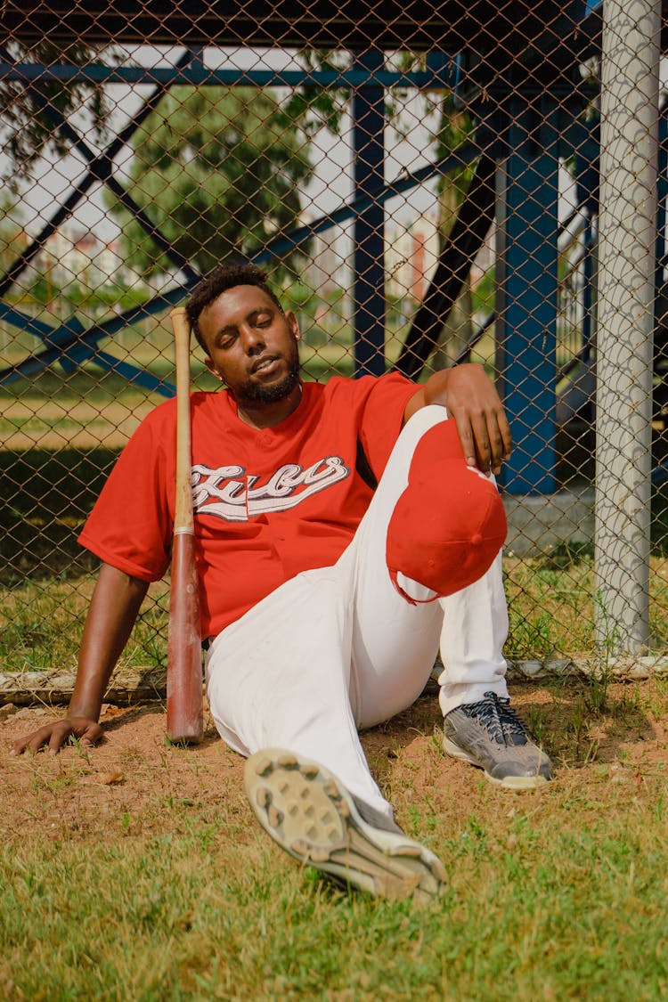 Man In Red And White Baseball Uniform Sitting On Ground