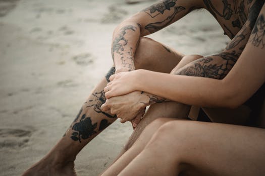 Close-up of a tattooed couple holding hands on a sandy beach, emphasizing intimacy.