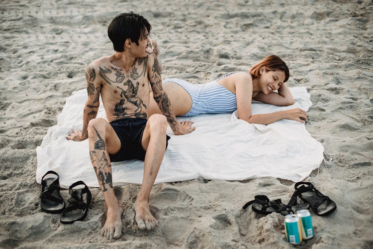 Tattooed Man Sitting On A Beach Towel Beside Woman Lying In Bikini