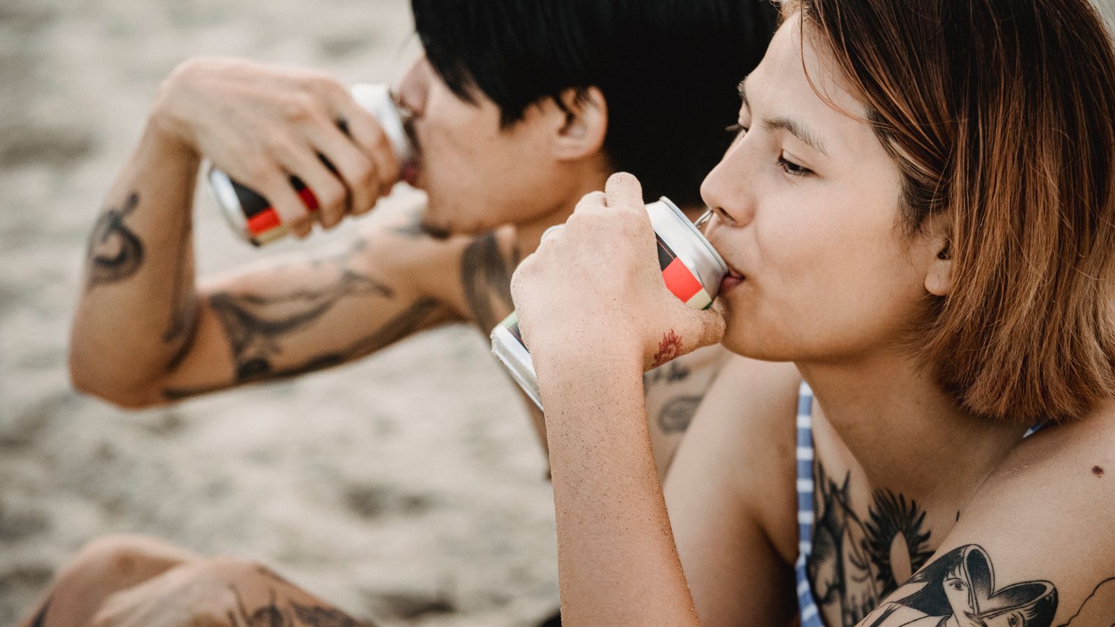 Two tattooed friends enjoying canned drinks while relaxing at the beach in summer.
