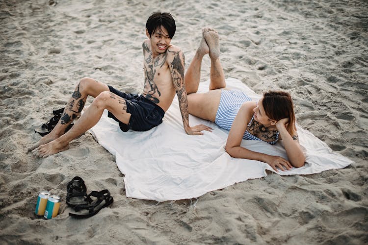 Tattooed Man Sitting Beside Woman Sunbathing On Beach Towel