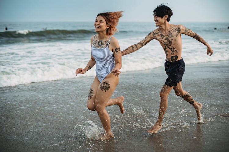 Tattooed Man Chasing Woman In Stripe Swimsuit Running On Beach