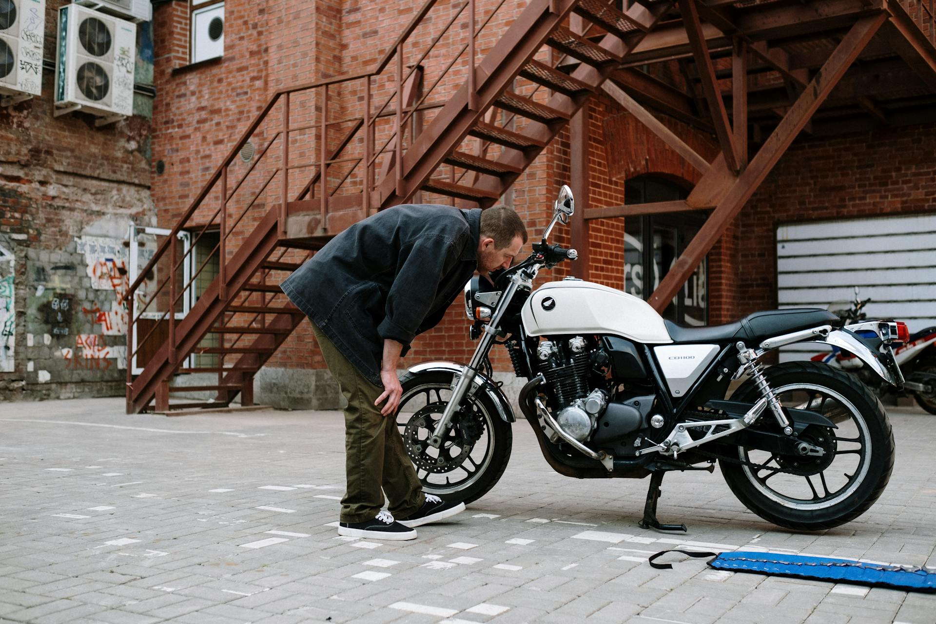 A man inspects a white motorcycle outdoors near a brick building with metal stairs.