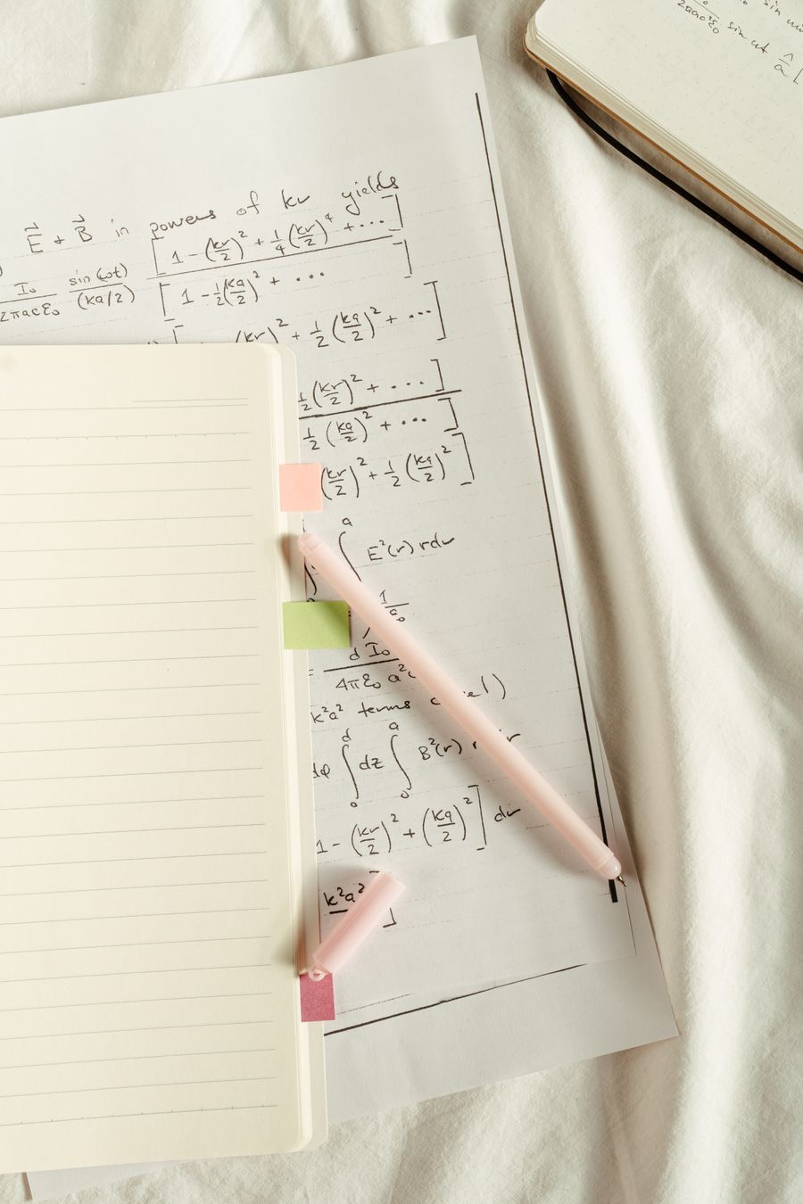 Notebook of handwritten mathematical formulas with coloured sticky notes and a pen