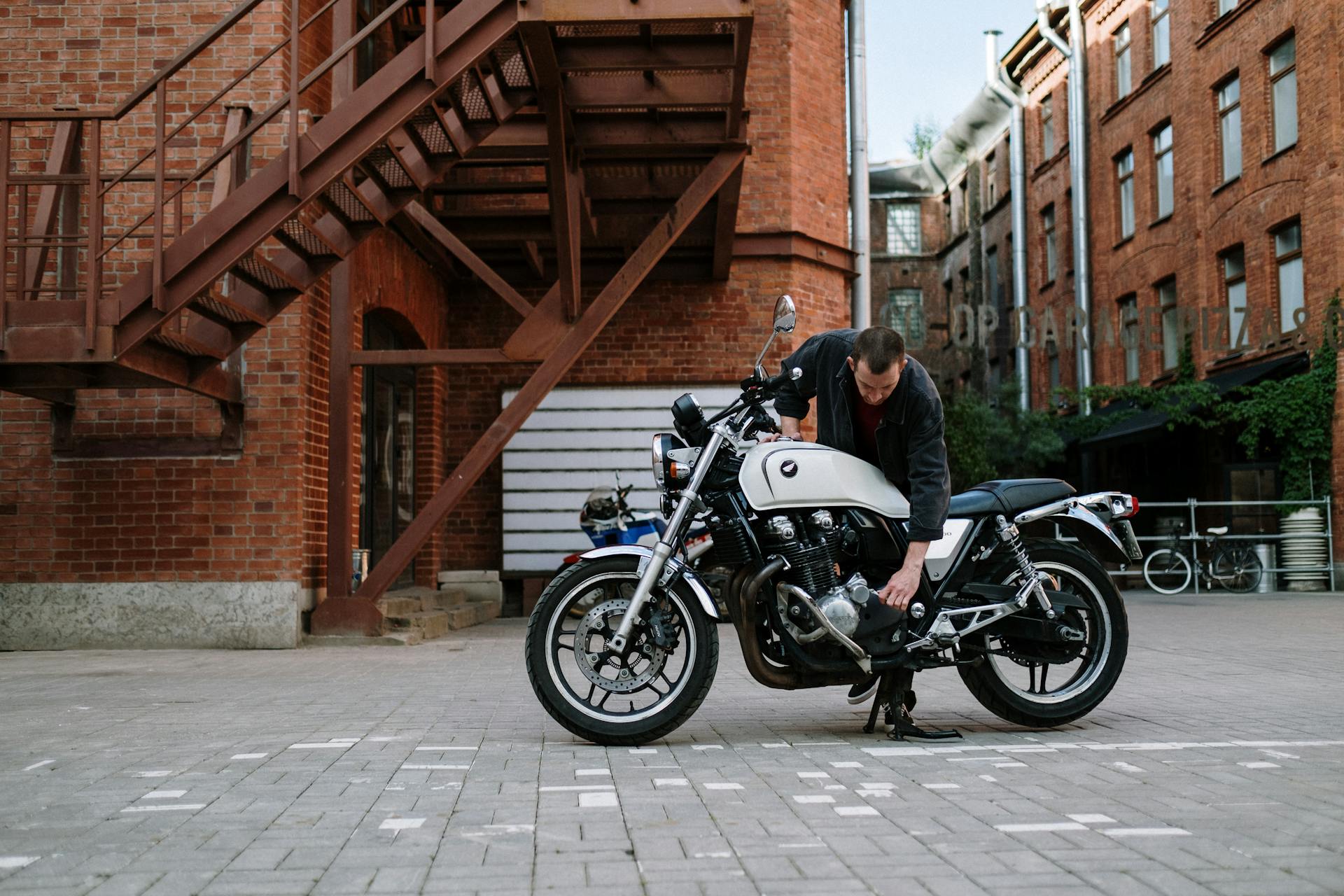 A man repairs a white motorcycle outside brick buildings, highlighting urban life and motorcycle maintenance.