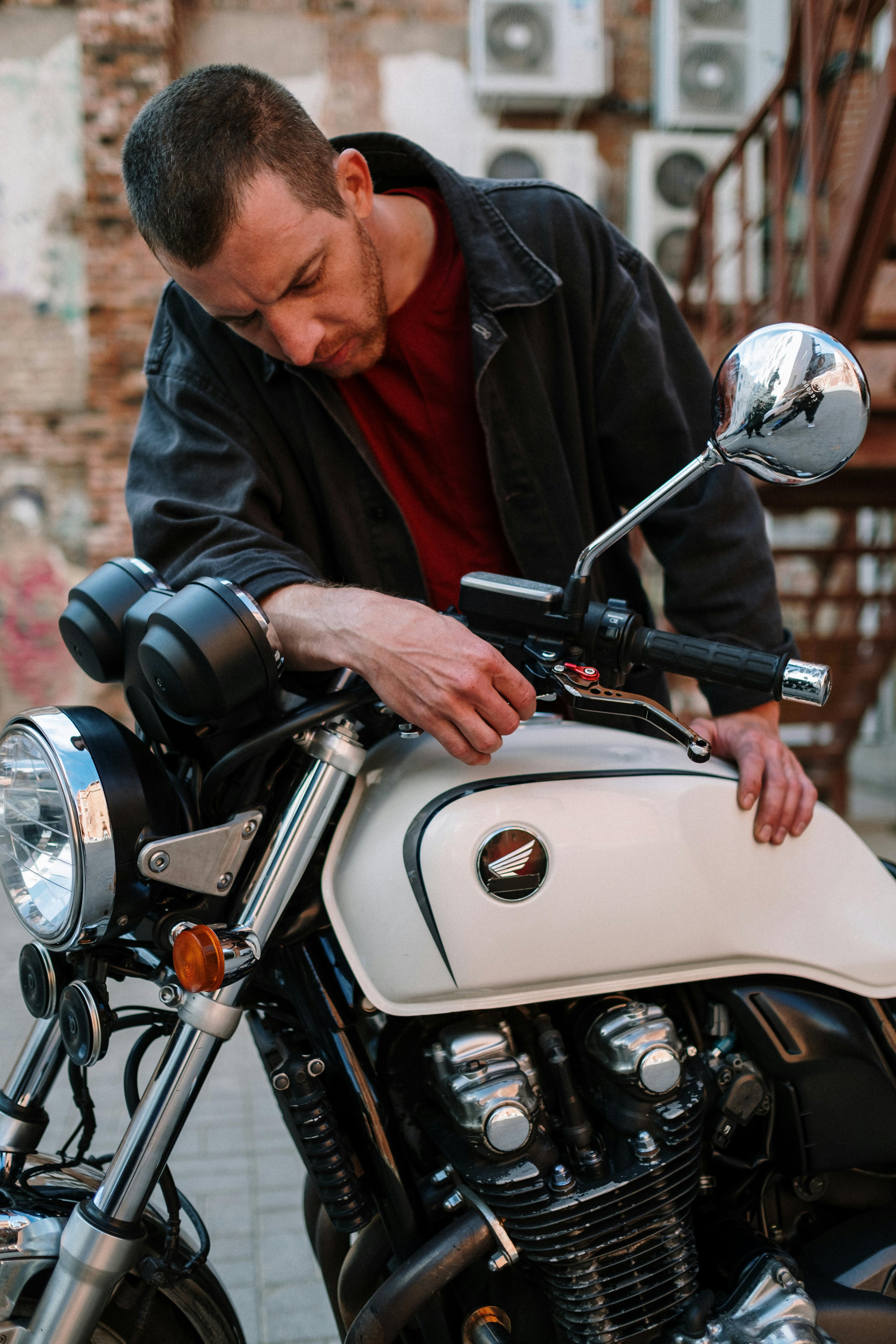 An adult male mechanic performs maintenance on a classic motorcycle outdoors.