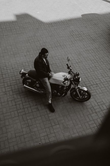 A stylish man sitting on a motorcycle in an urban area, captured in monochrome from a high angle.