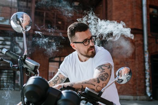 一名留着纹身、蓄着时尚胡须的男子骑着摩托车，在户外吐着烟