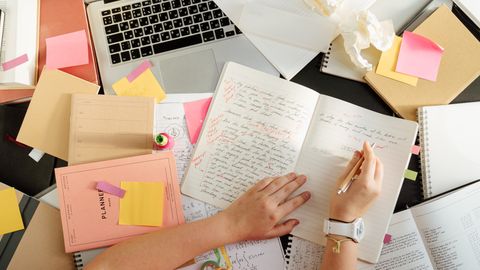 Overhead shot of a person organizing notes and study materials on a cluttered desk with a laptop.