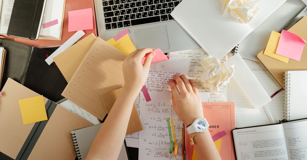 Student working on a laptop in a library, surrounded by sticky notes and a smartphone face-down