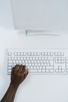 Close-up of a hand typing on a white computer keyboard against a white surface.