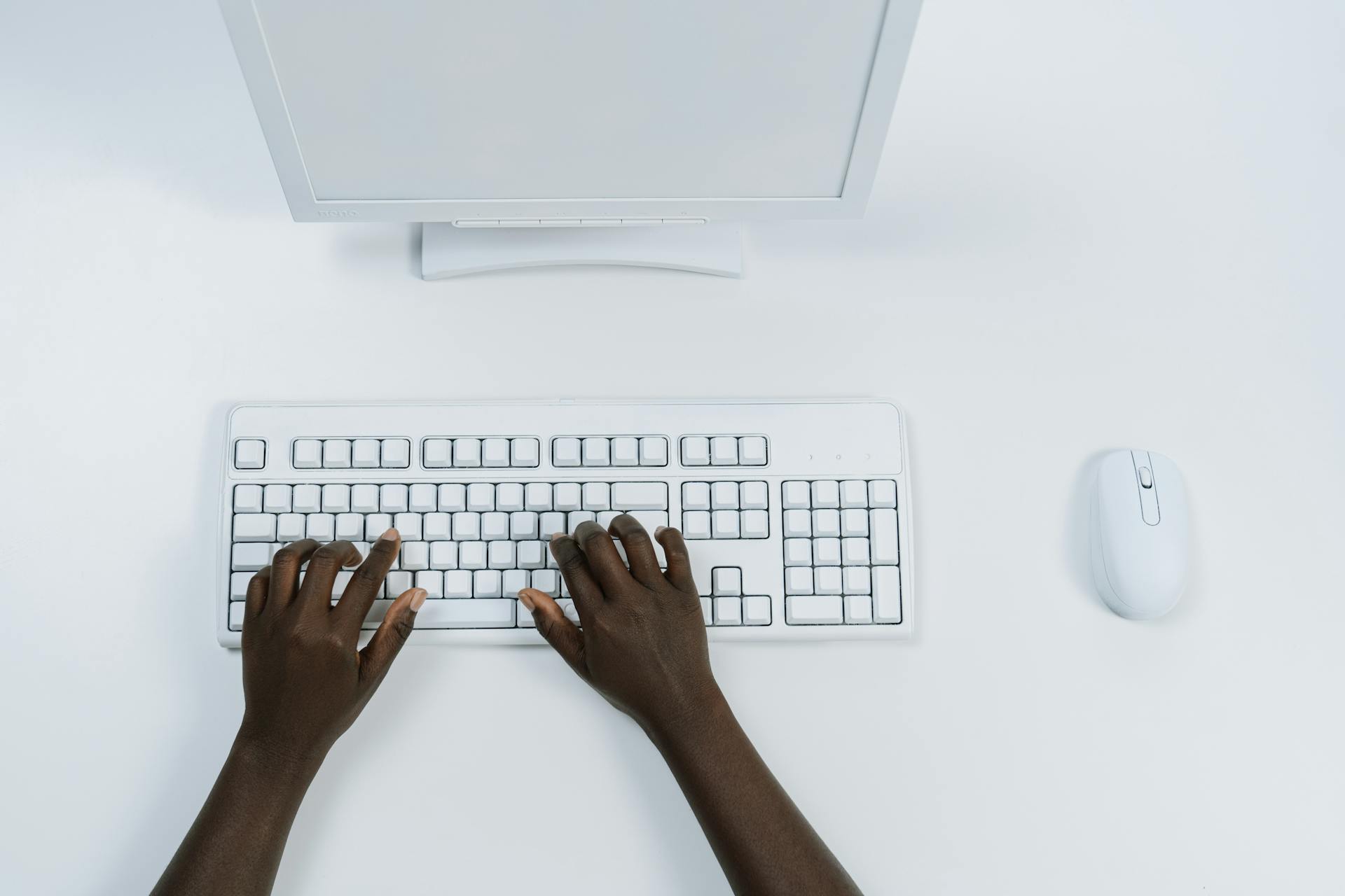 Overhead view of hands typing on white computer setup with mouse.