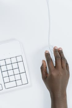 Close-up of a hand using a computer mouse next to a keyboard on a white desk, showcasing minimalist design.