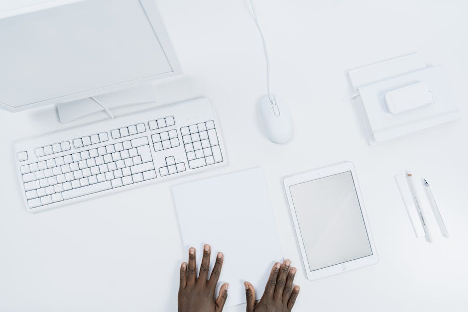 Top view of a minimalist white office workspace with gadgets and a person's hands.