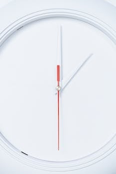 这是一款时尚的白色壁挂时钟特写，采用极简主义设计，时钟指针为红色和白色