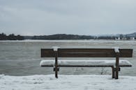 Snow Covered Bench in Front of Body of Wa Ter