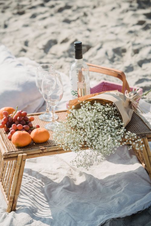 Free Elegant beach picnic setup with wine, grapes, flowers, and a wicker tray on sand. Stock Photo