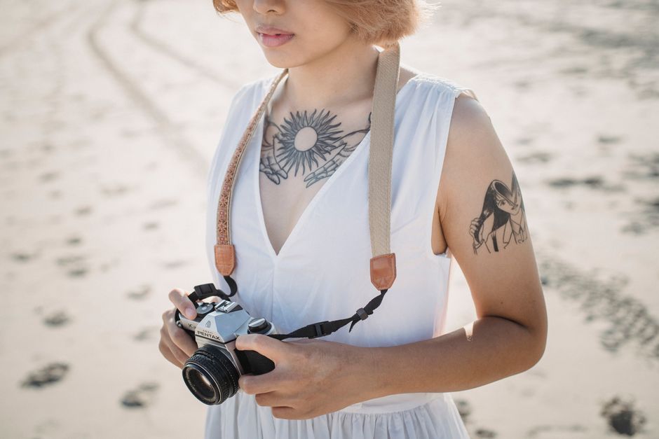 A woman with tattoos holding a vintage camera on the sandy beach.