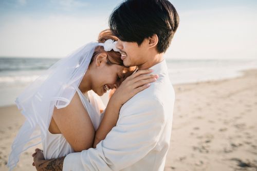 Free Happy couple embracing on a sandy beach during their wedding day, capturing a joyful moment. Stock Photo