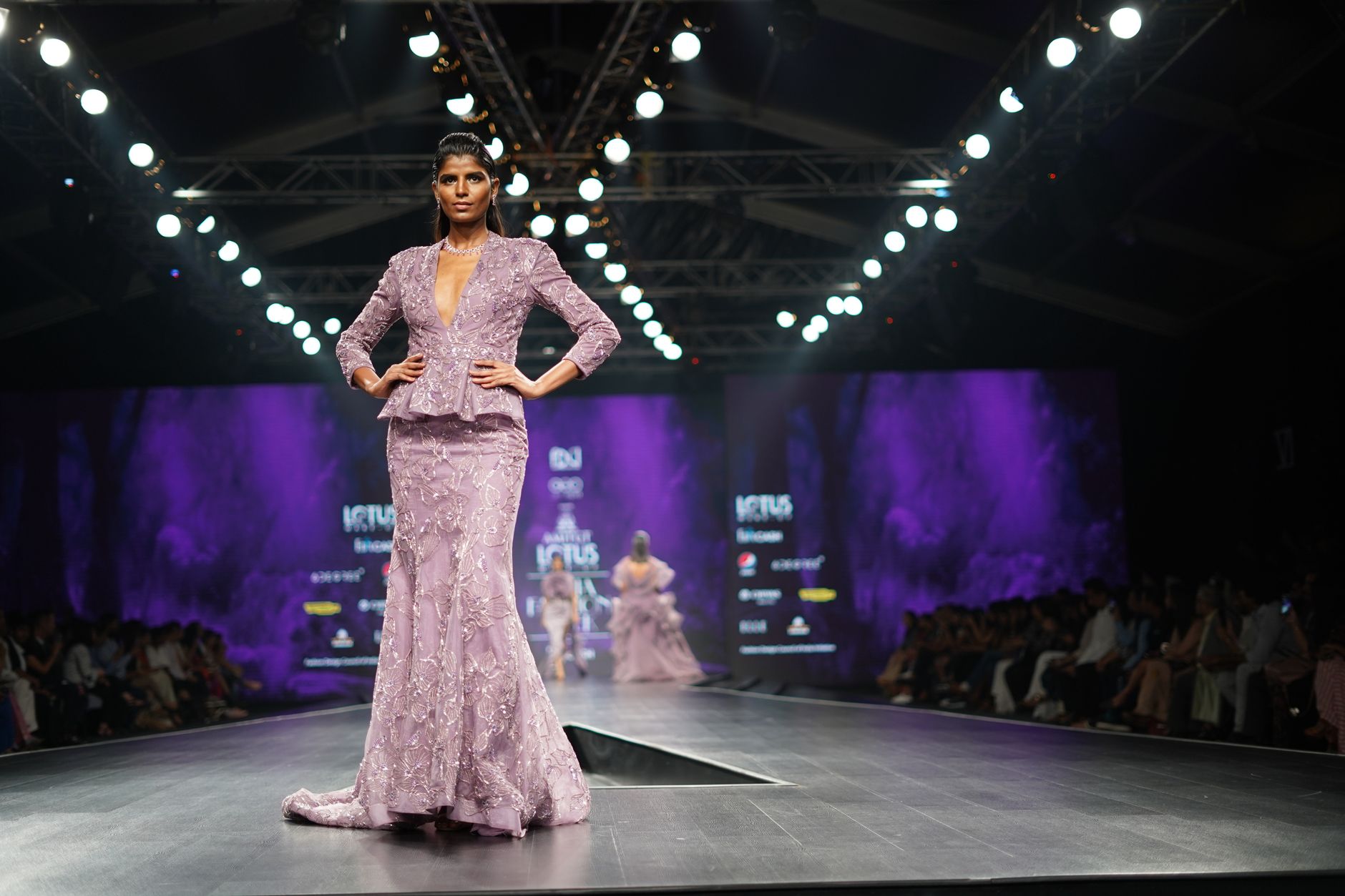 A beautiful model in a lavender dress gracefully poses on the runway at a fashion show.