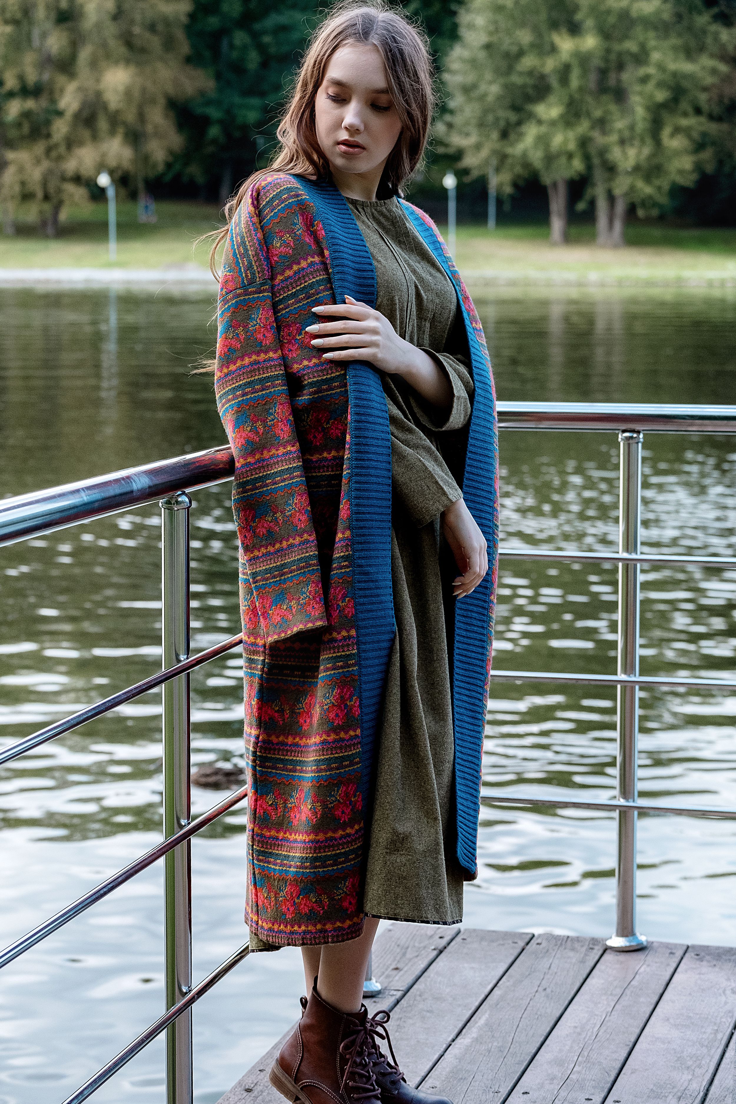 Attractive female wearing trendy ornamental coat standing on peaceful pond pier in green park and looking down thoughtfully