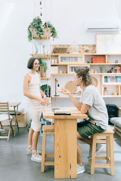 Free Two colleagues engage in a casual meeting in a modern office setting with plants and bookshelves. Stock Photo