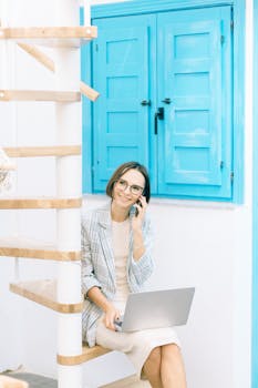 Professional woman in business attire using phone and laptop at home office space.