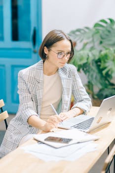 Professional woman in a blazer working on a laptop at a wooden desk with plants and blue decor.